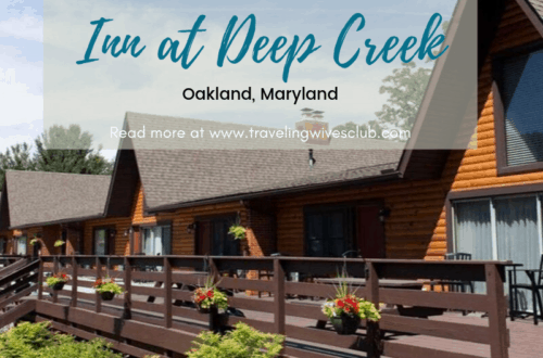 Brown sided lodging with deck. Small flowers hanging off of deck and green plantings in front.